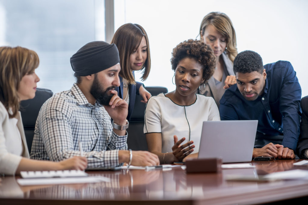 A group of business people gathered around a laptop in a modern office and discussing what they see.